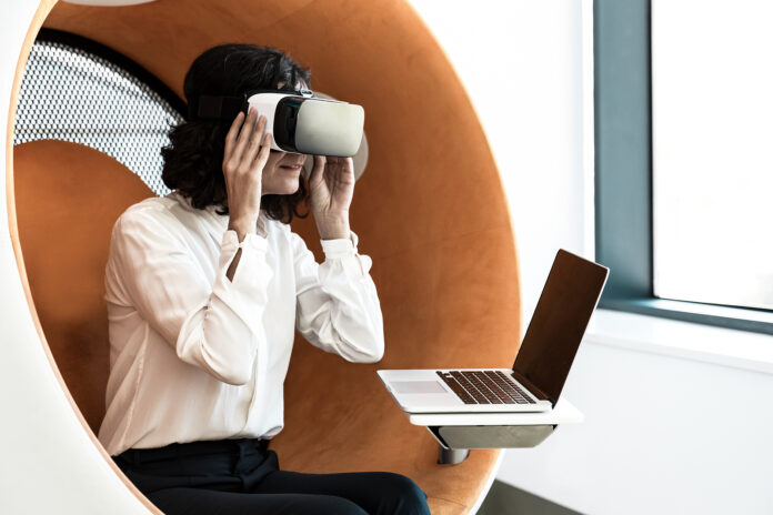 Woman watching virtual presentation with VR goggles and a laptop.