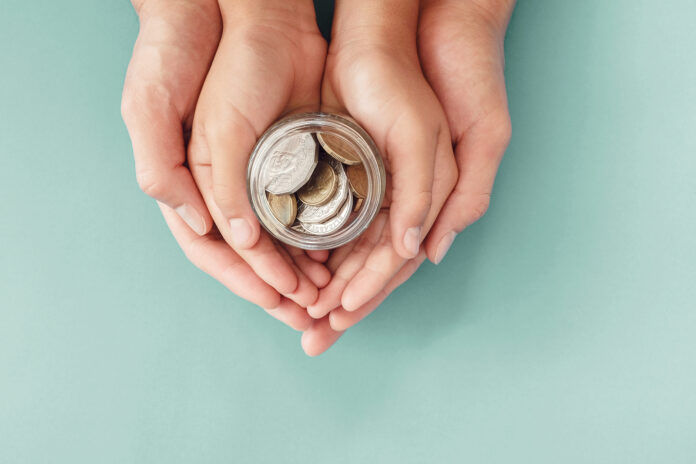 Child and parent hands holding money jar filled with coins.